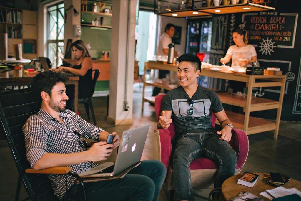 Two young men sitting in a cafe, one with a laptop and the other holding a phone, both smiling and engaging in a conversation.