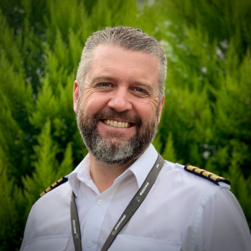 Smiling male pilot in a white shirt with epaulettes and a lanyard, standing in front of green foliage.